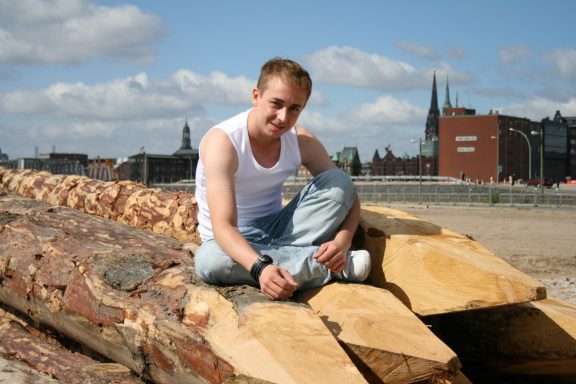 Junger Mann sitzt lachend auf großen Holzstämmen mit Stadt im Hintergrund.