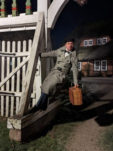Mann im Mantel mit einem Koffer, sitzt auf einer Holzplattform bei Nacht.